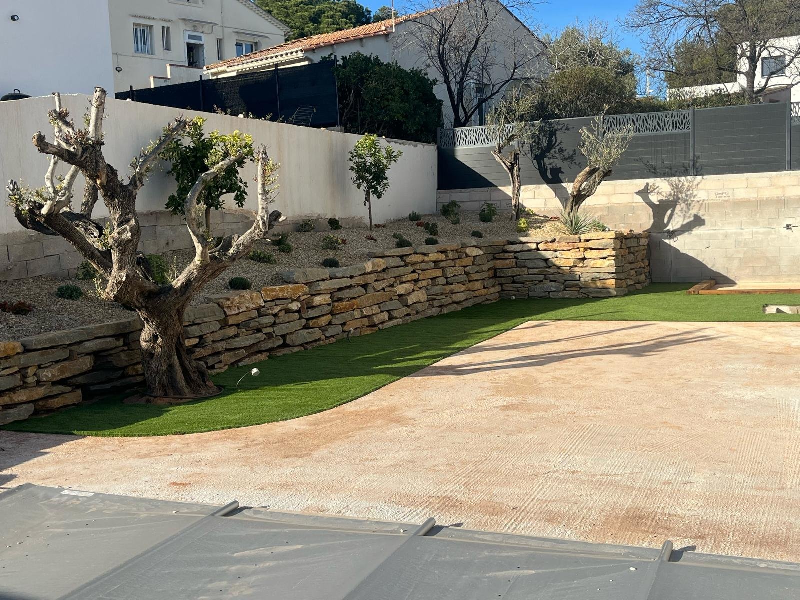 Un jardin aménagé avec des murs de soutènement en pierres sèches, des plantations, du gazon synthétique et une grande zone en béton clair au premier plan.