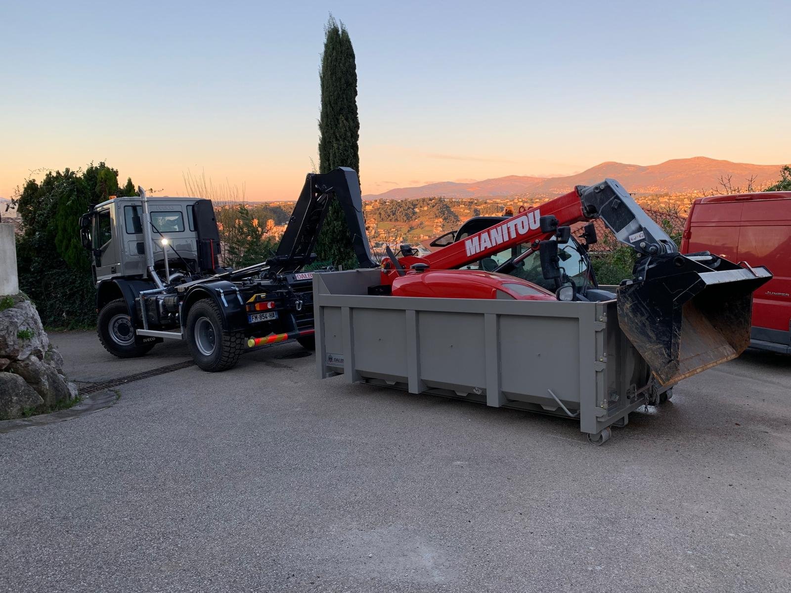 Un camion Iveco gris avec un bras de levage transporte un engin Manitou rouge dans une benne de chantier, sur une allée, avec un coucher de soleil et des collines en arrière-plan.