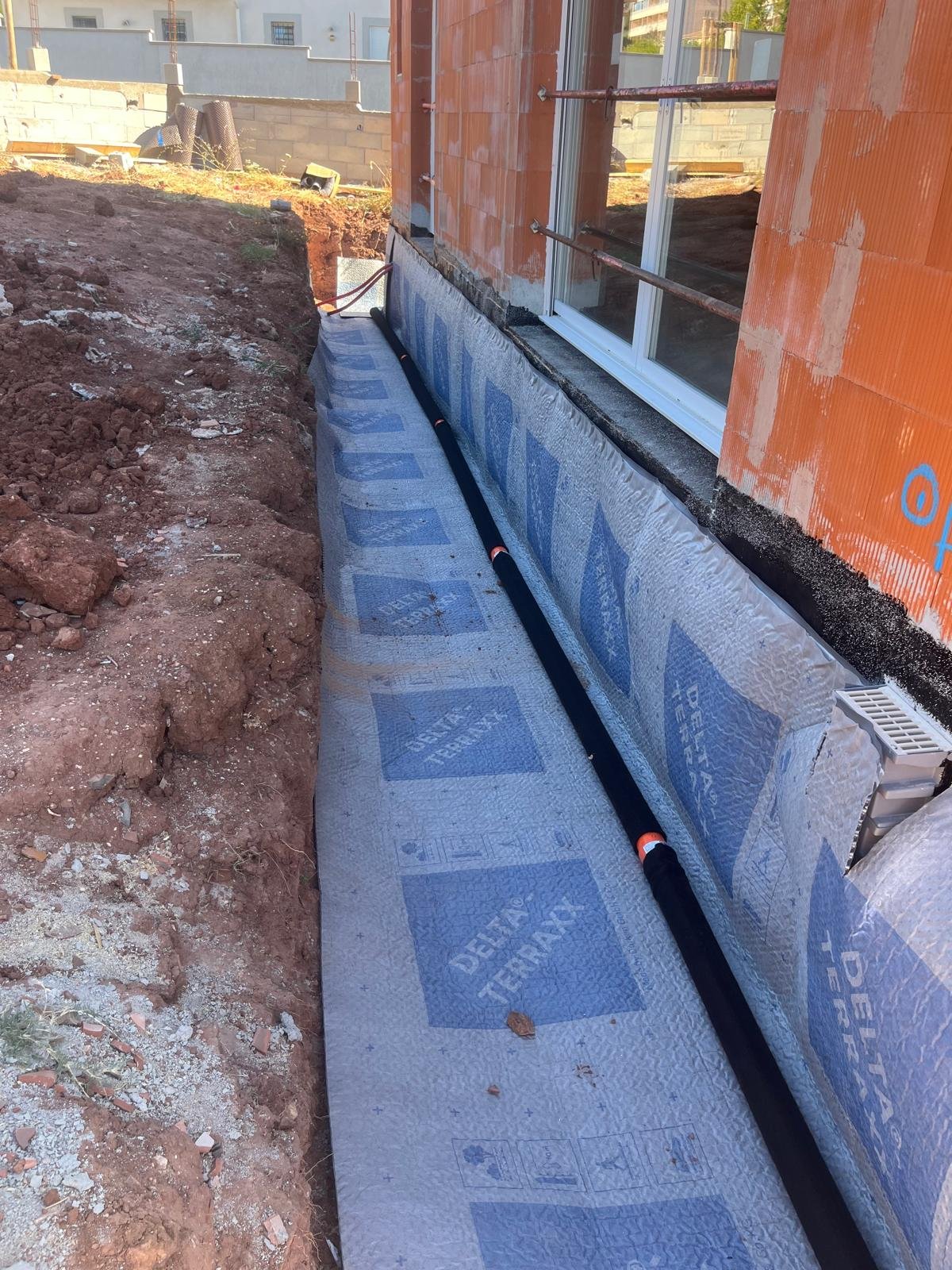 Une tranchée de drainage est creusée le long d'une fondation de maison, recouverte d'une membrane d'étanchéité bleue DELTA TERRAXX et d'un tuyau de drainage noir.