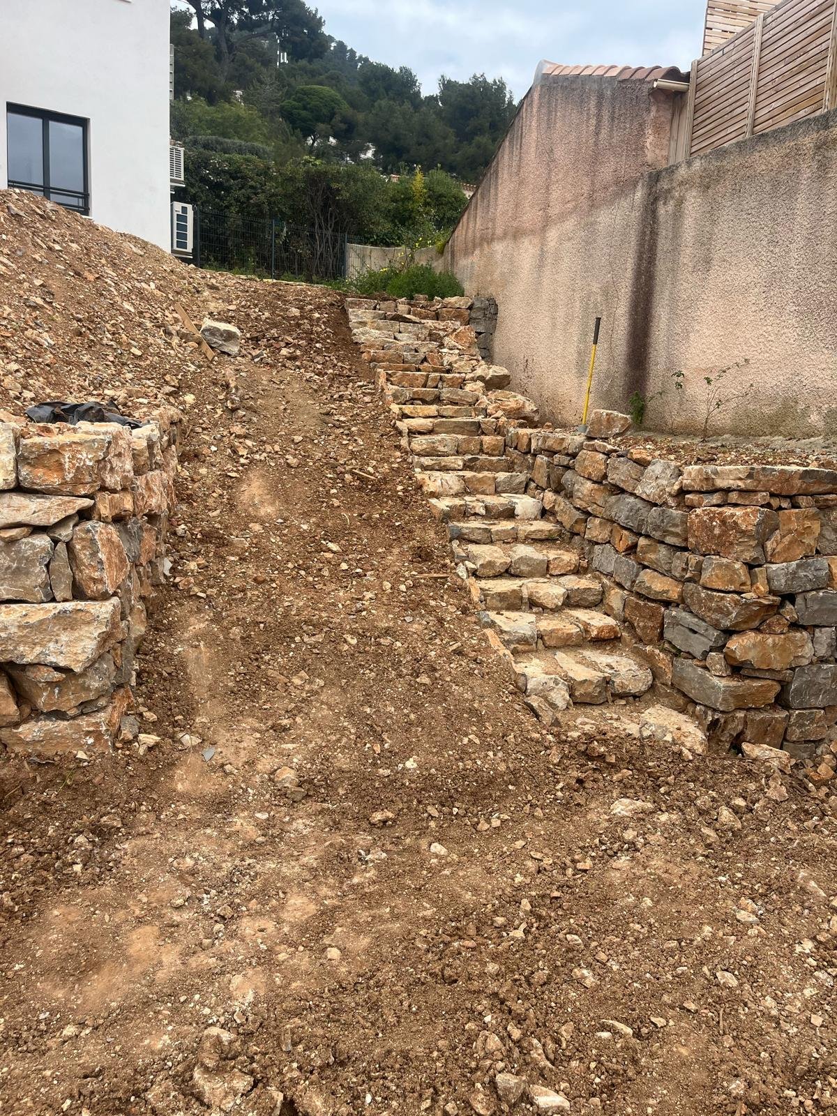 Image d'un chantier de terrassement avec un escalier et des murs de soutènement en pierre sèche sur un terrain en pente, devant une maison blanche et un mur de clôture.