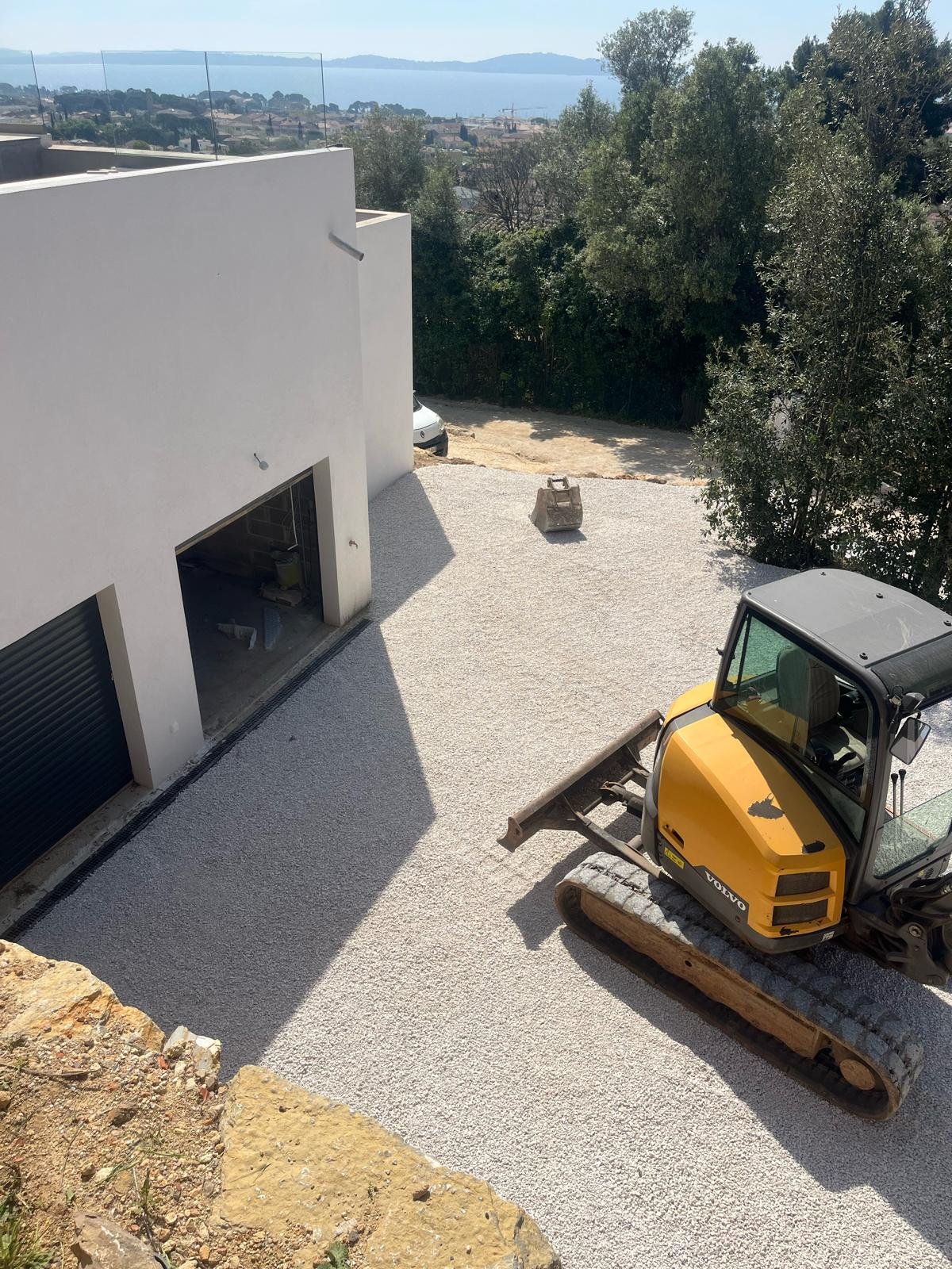 Une excavatrice Volvo jaune est garée sur une allée de gravier devant une villa moderne avec des garages noirs, avec la mer Méditerranée et des collines visibles à l'arrière-plan.