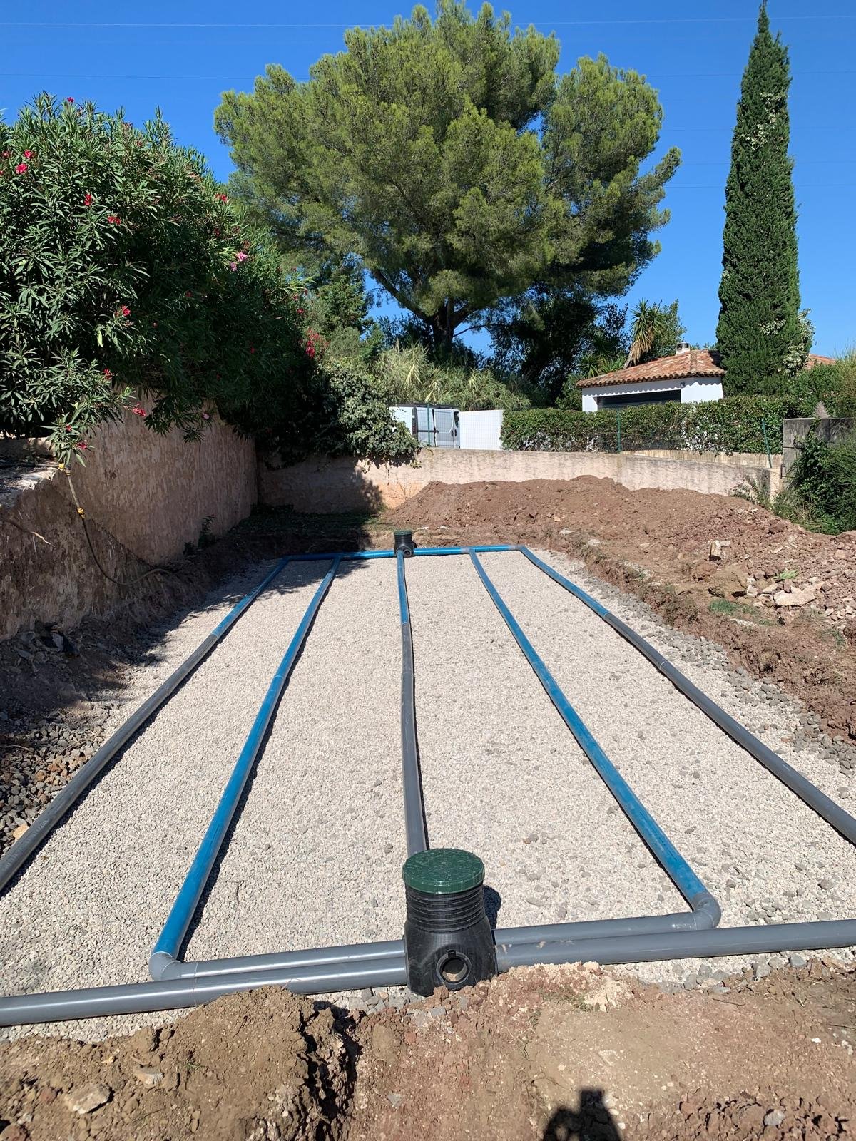 Installation d'un système d'assainissement non collectif avec des tuyaux de drainage bleus et gris posés sur un lit de gravier dans une tranchée.