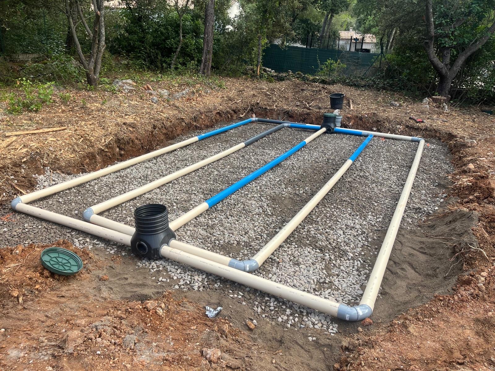 Un système de champ d'épandage pour fosse septique est en cours d'installation, montrant des tuyaux beige et bleus posés sur un lit de gravier dans une excavation de terre.