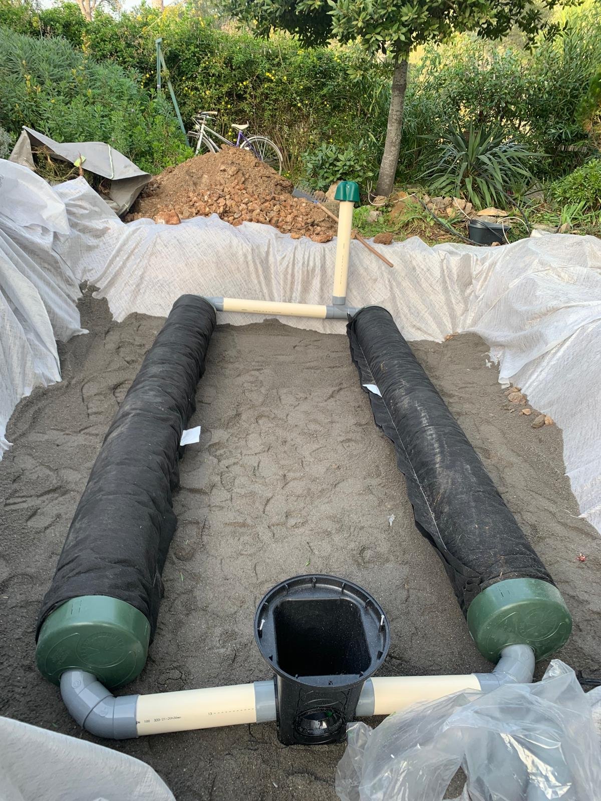 Installation d'un système de drainage avec deux tuyaux noirs enveloppés de géotextile, posés sur un lit de sable et raccordés à un regard central, dans une zone de chantier paysager.