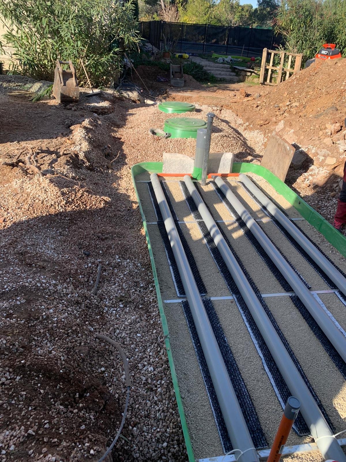 Installation d'un filtre à sable compact avec des tuyaux gris posés sur un lit de gravier, en cours sur un chantier de terrassement ensoleillé.