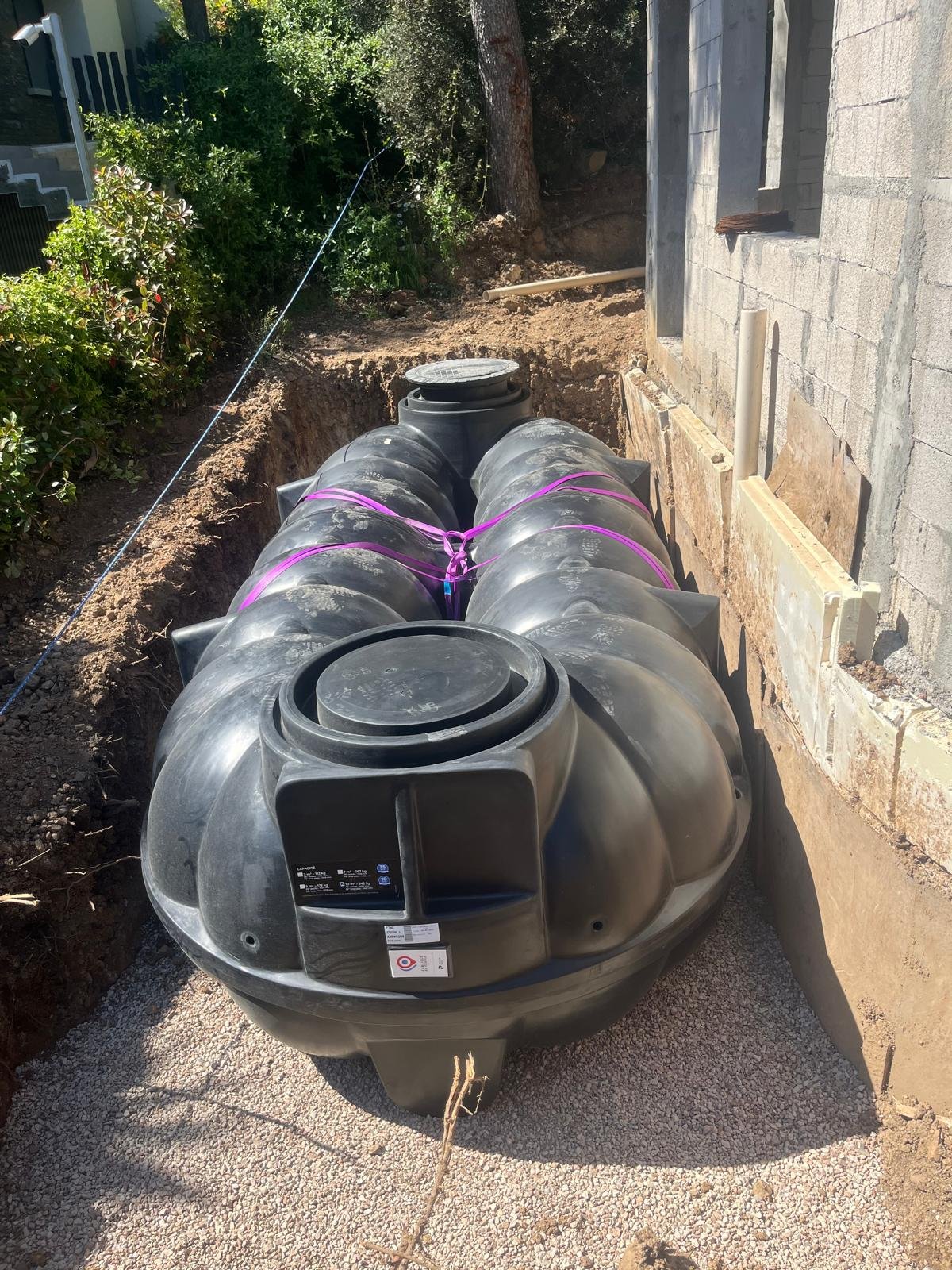 Deux grands réservoirs noirs interconnectés sont installés dans une tranchée de gravier profonde à côté d'un mur en parpaings, avec des sangles violettes les fixant.