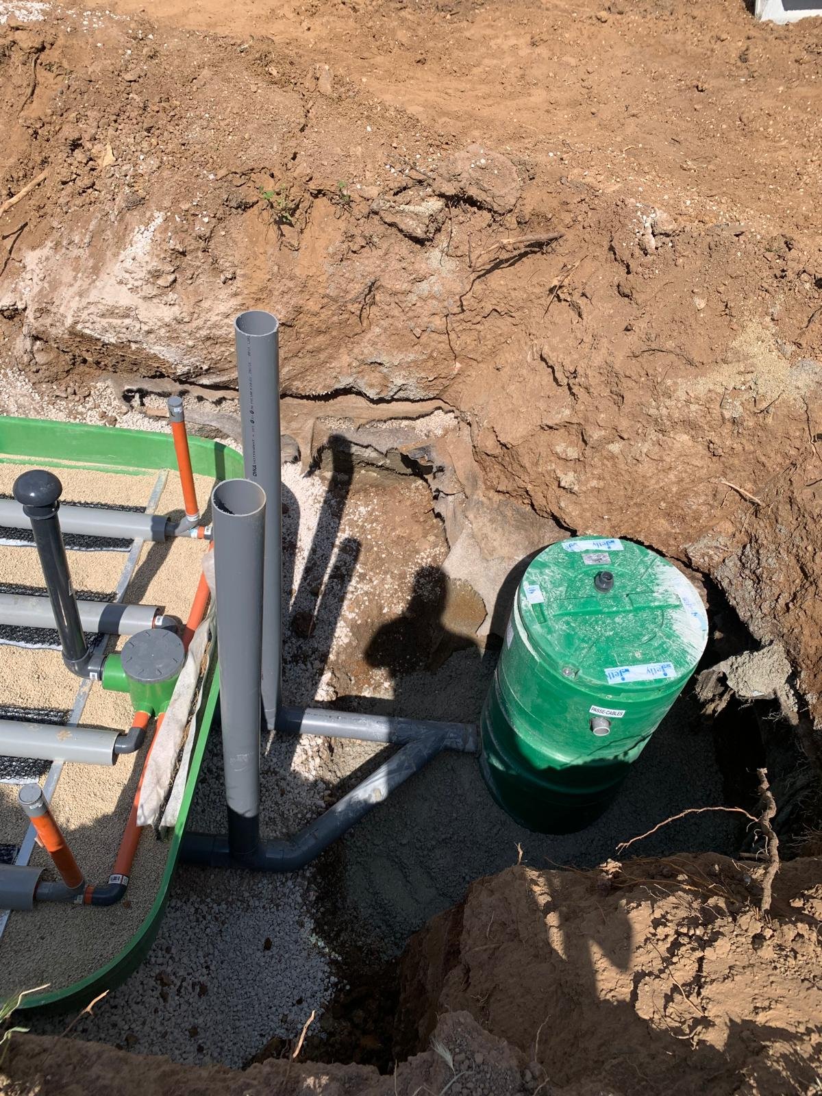 Image d'une fosse septique verte et de tuyaux gris et oranges en cours d'installation dans une tranchée de terre meuble sur un chantier de terrassement.