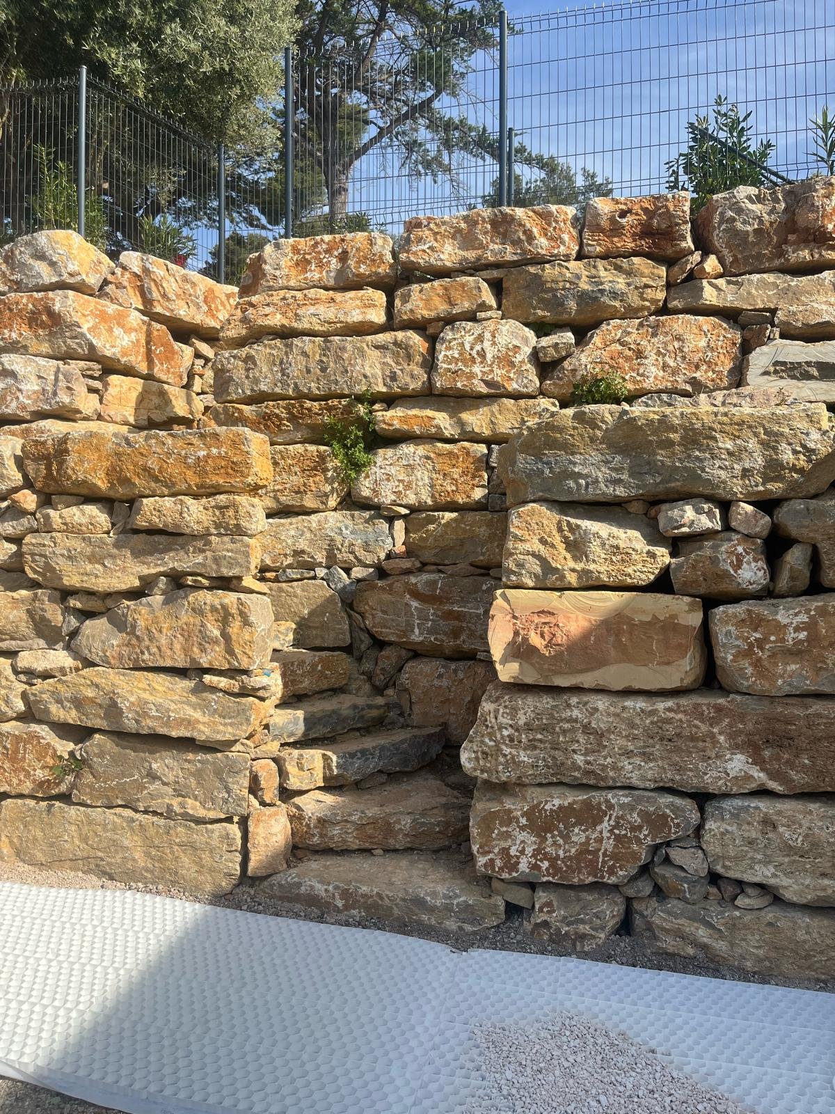 Mur de soutènement en pierres sèches ocre, avec une clôture métallique, des arbres et une grille de stabilisation au sol.