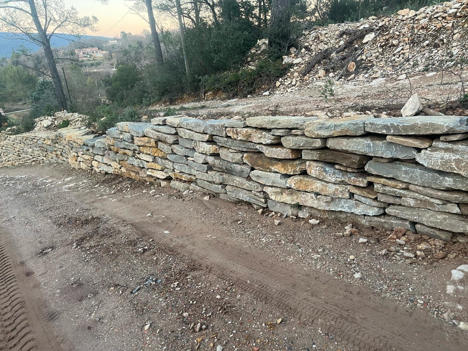 Vue latérale d'un mur en pierres sèches brun et gris, longeant un chemin de terre avec des traces de pneus, et un paysage vallonné boisé en arrière-plan sous un ciel clair.