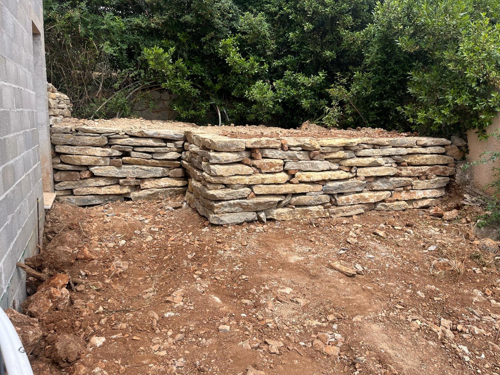 Deux murets en pierres sèches avec un remblai de terre rouge en chantier, devant une végétation dense, illustrant un aménagement paysager.