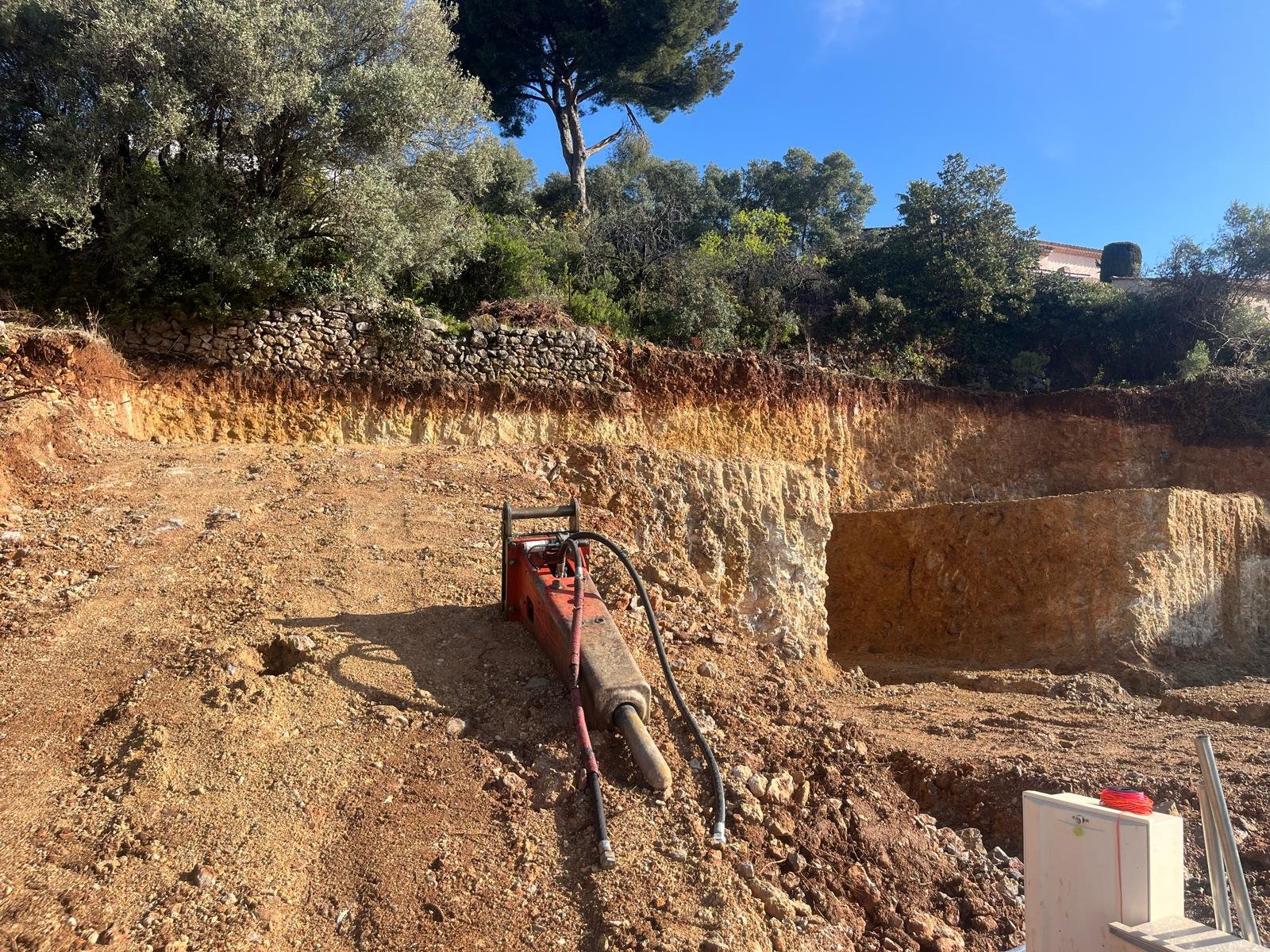 Un brise-roche hydraulique rouge et gris repose sur le sol d'un chantier de terrassement, devant une paroi rocheuse récemment excavée, sous un ciel bleu.