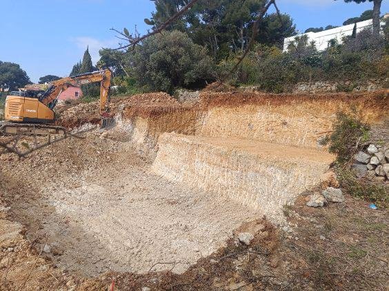 Une pelle mécanique orange effectue une grande excavation sur un chantier de terrassement, exposant des couches de terre et de roche sous un ciel bleu.