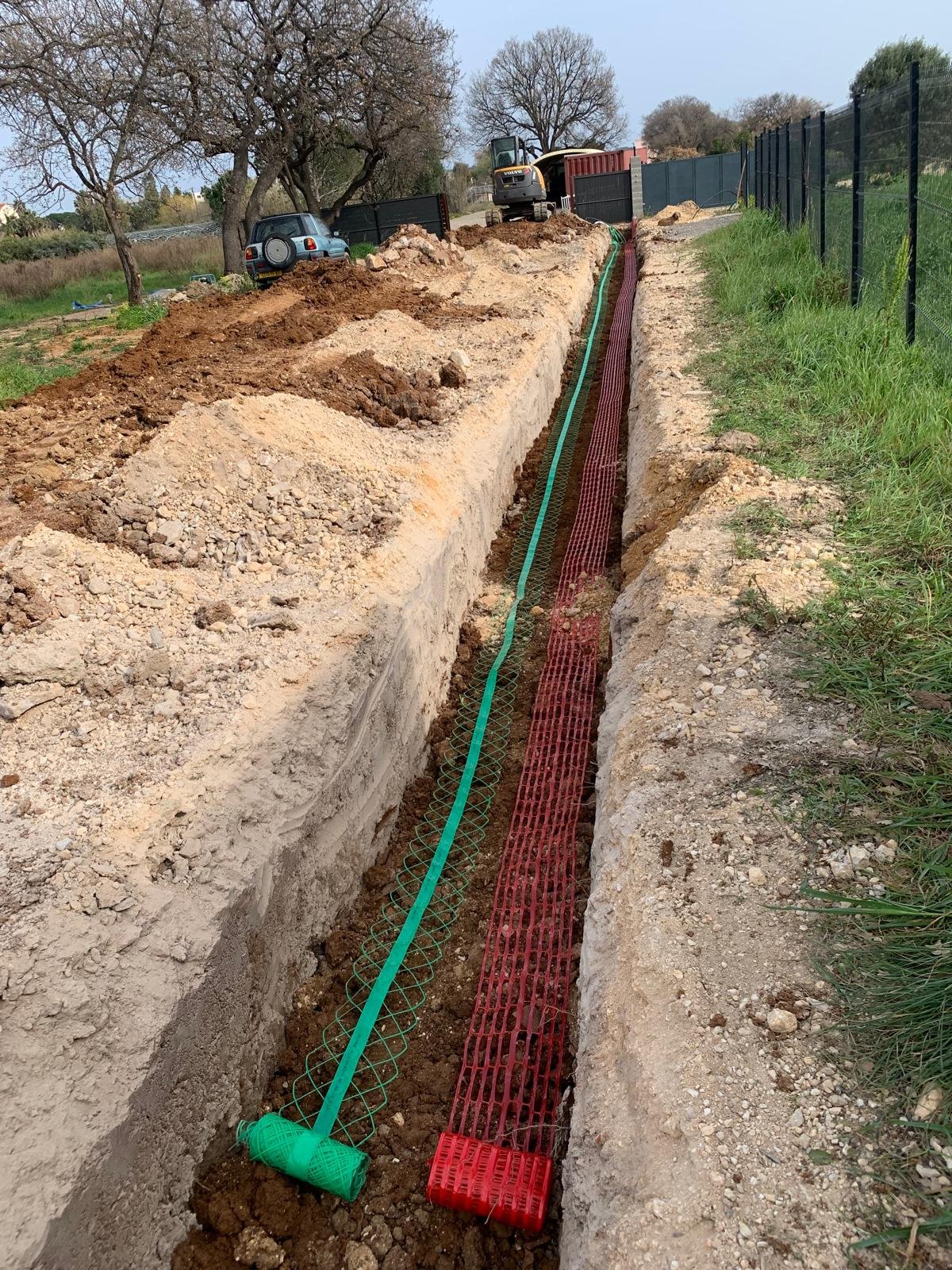 Une longue tranchée creusée expose des filets de signalisation verts et rouges installés pour protéger des conduits souterrains, avec un engin de chantier et une voiture visible en arrière-plan.