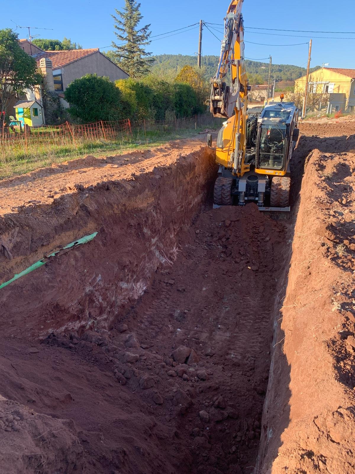 Une pelleteuse jaune creuse une longue tranchée dans de la terre rouge, avec un tuyau vert visible sur le côté et des maisons en arrière-plan.